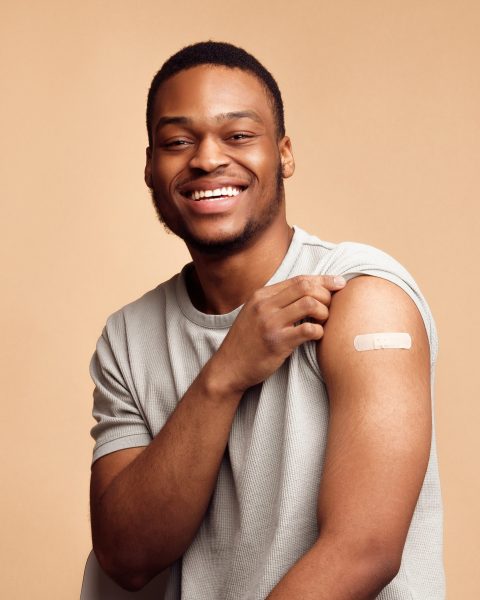 Covid-19 Vaccination. Happy Vaccinated African Man Showing Arm With Plaster After Coronavirus Vaccine Injection Sitting Over Beige Studio Background, Smiling To Camera. Flu Shot, Virus Prevention